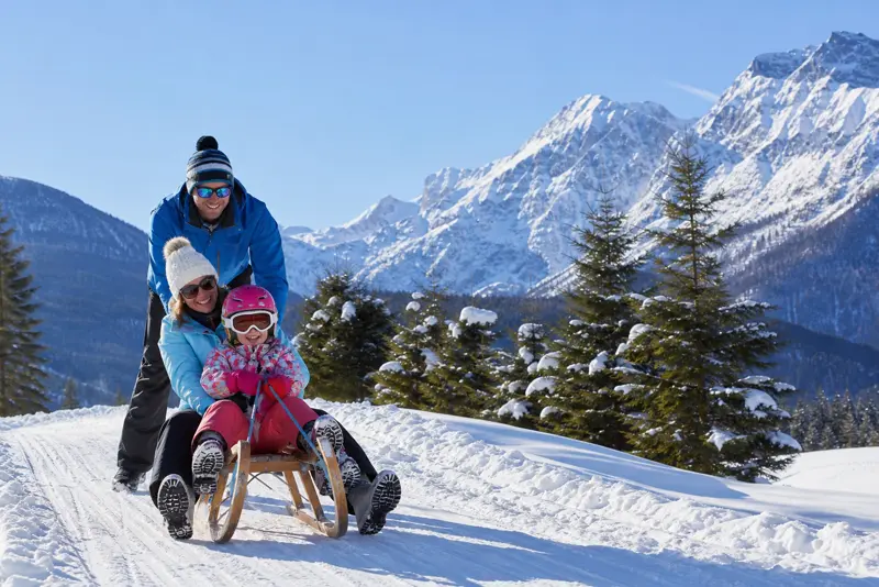 Familie beim Rodeln vor verschneiten Bergen in einem Skiort